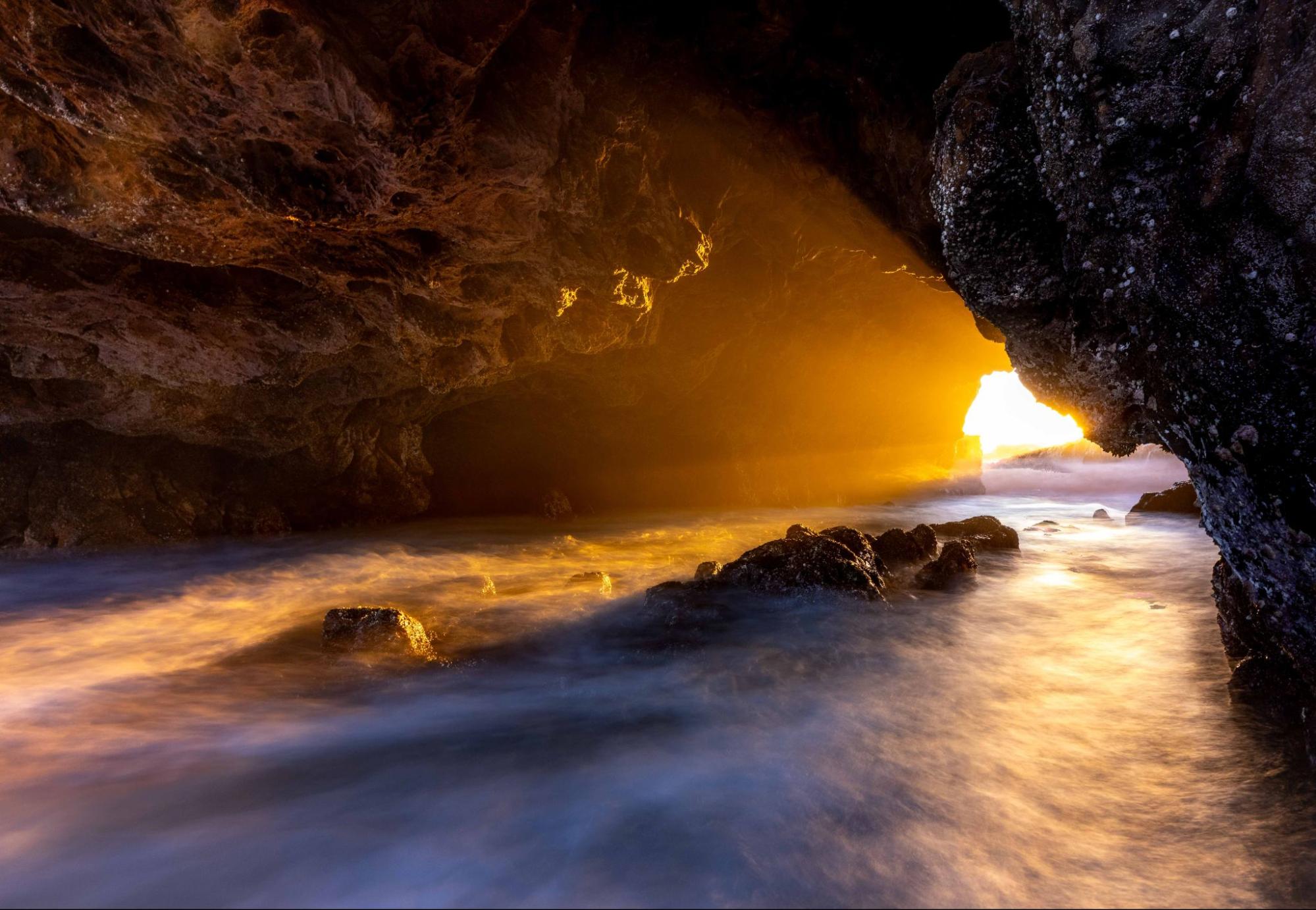 Sunset illuminating a sea cave entrance; ocean water flows over rocks, glowing orange. Photo by Tim Truby.