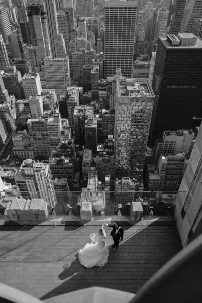 Black and white photo: a bride and groom dance on a rooftop overlooking a cityscape