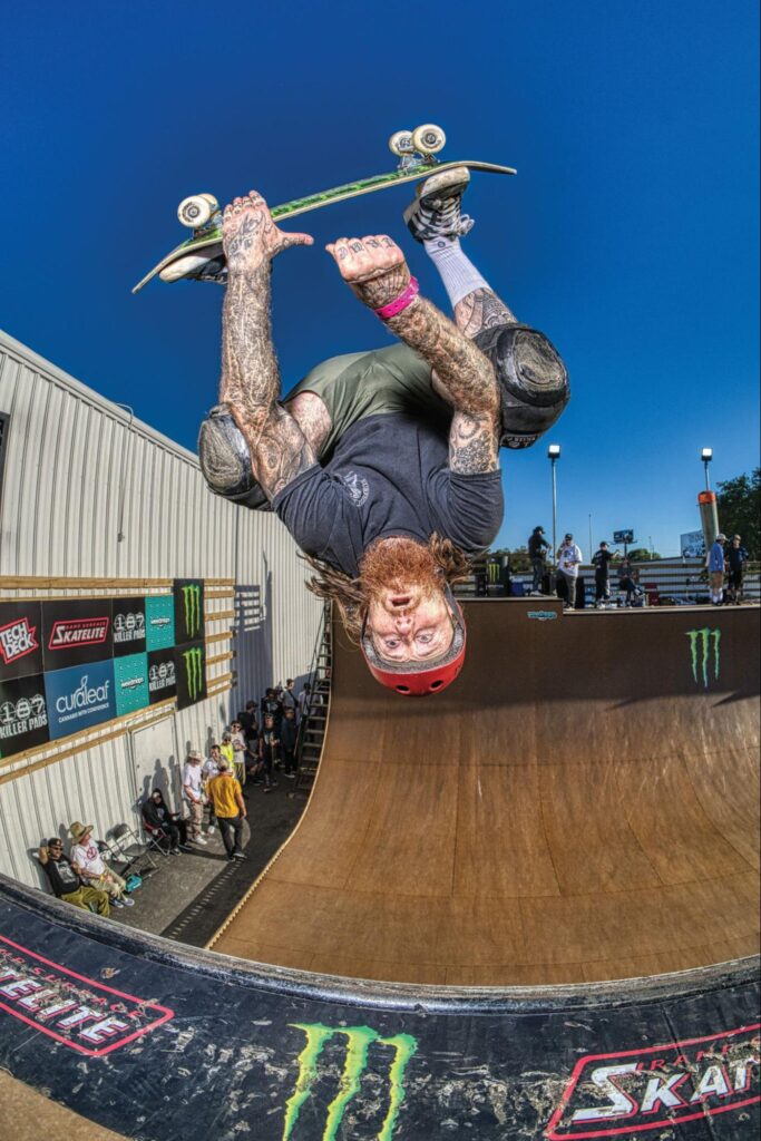 Tattooed skateboarder upside down, performing a trick in a ramp, spectators watching