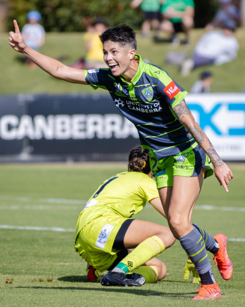 Jubilant female soccer player in lime green and grey uniform celebrates, arm raised. Photograph by Denis Hawkins.