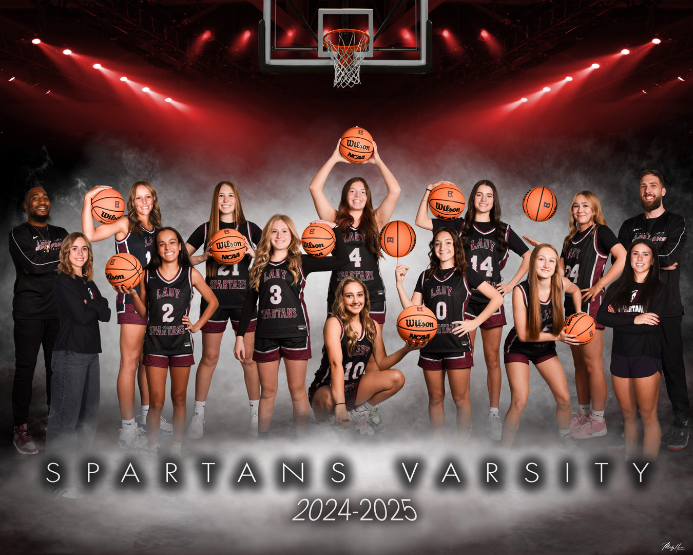 The image shows the 2024-2025 Spartans Varsity girls' basketball team, posing with basketballs in front of a backdrop featuring a basketball hoop and red stage lighting. The team members are wearing black and maroon uniforms with "Lady Spartans" and their numbers displayed. Image by Misty Huss Photography