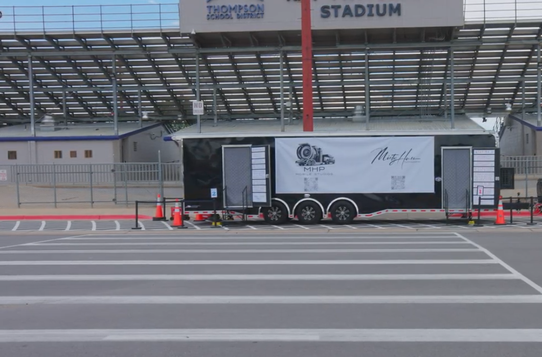 A black mobile photography studio trailer with the logo "MHP Mobile Studios" is parked in front of a stadium. The trailer has two open doors and a white banner with the studio's name and logo.