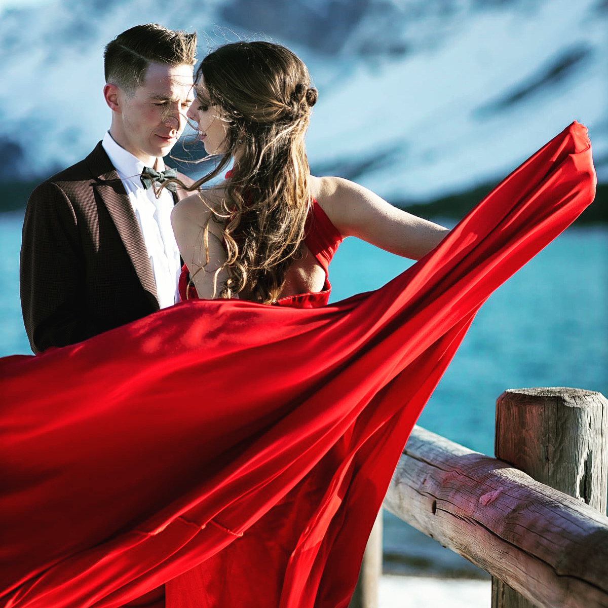 A couple poses romantically by a lake, with the woman in a flowing red dress that billows in the wind. The man is in a suit and bow tie, and snow-capped mountains are visible in the background. Photo by William Orsua