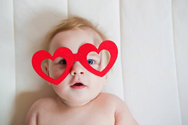 A baby with blonde hair and blue eyes lies on a white surface, wearing oversized red heart-shaped glasses. The baby looks directly at the camera with a slightly open mouth.