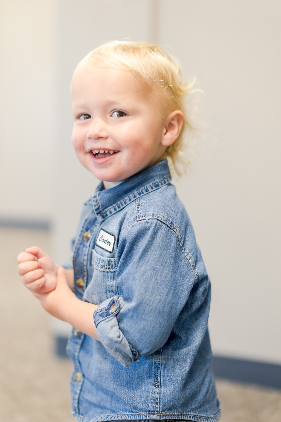 A young child with blonde hair and a denim shirt smiles at the camera. The shirt has a name tag that reads "Carter."