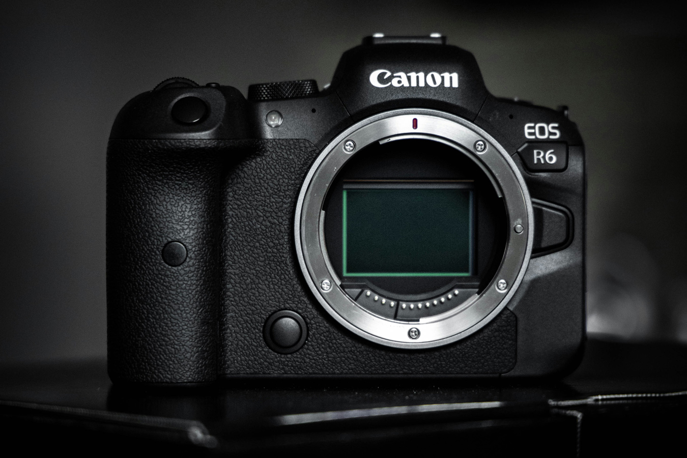 A close-up shot of a black Canon EOS R6 camera body, without a lens, reveals the sensor inside the lens mount. The camera is positioned on a dark, reflective surface.