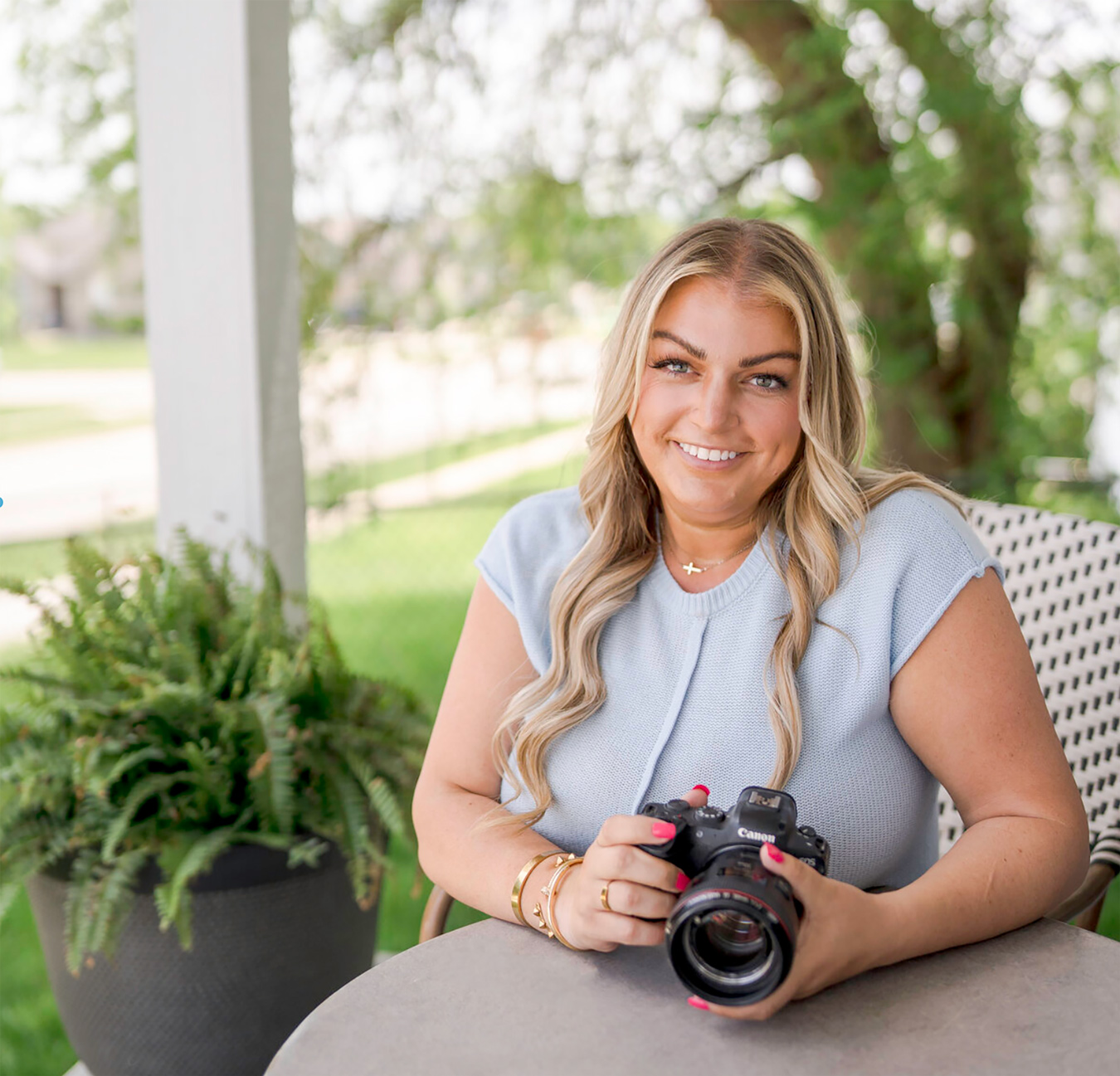 A smiling woman with blonde hair sits at a table outdoors, holding a Canon camera with a large lens. She wears a light blue top, gold jewelry, and has pink nail polish.