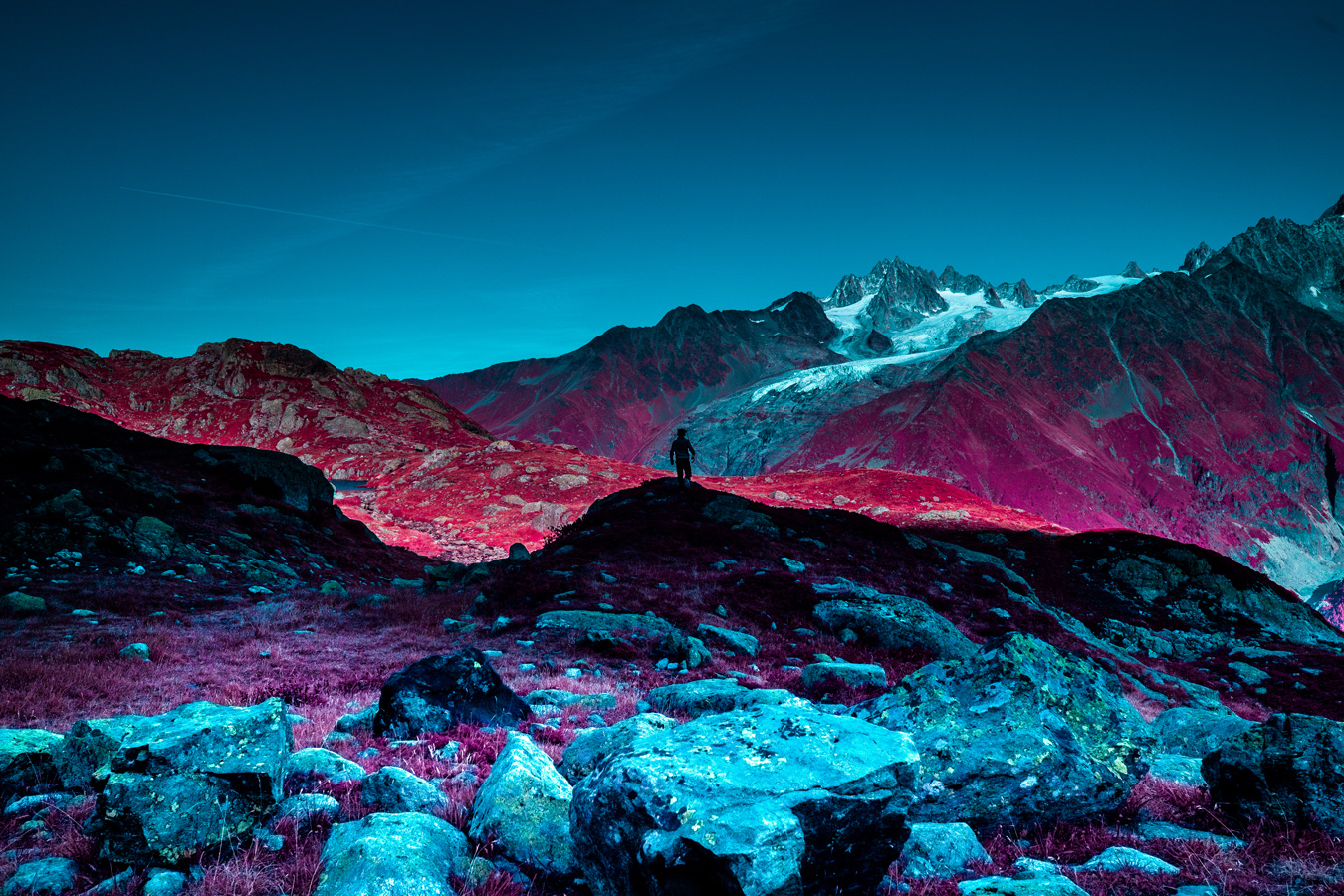 A lone figure stands atop a rocky hill in a surreal landscape of turquoise rocks, magenta grass, and red mountains, with snow-capped peaks in the background under a clear blue sky. The scene is bathed in an otherworldly light, creating a striking contrast of colors. Photo by Katie Farr.