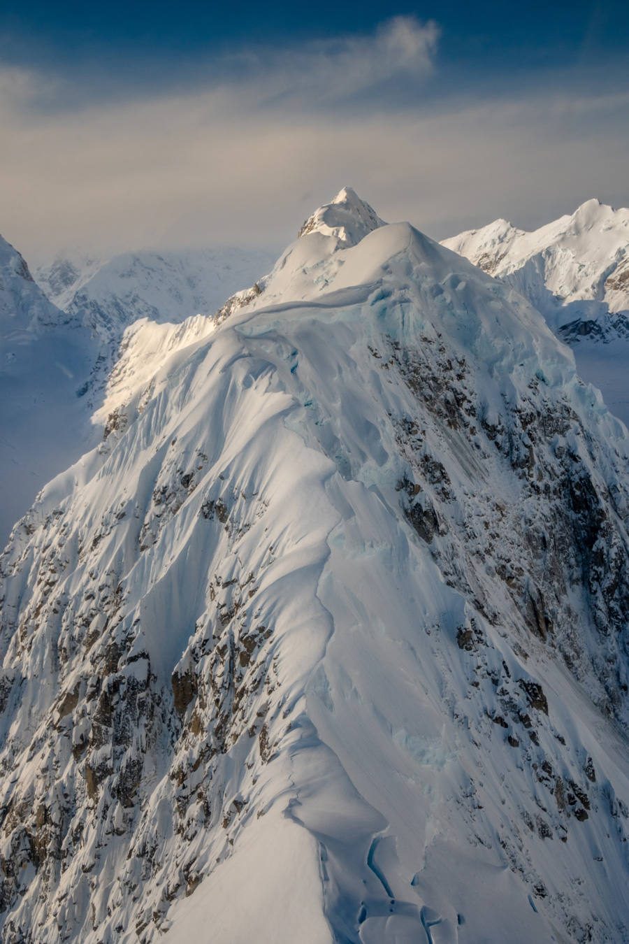 A high-angle shot captures a snow-covered mountain peak with rocky outcrops, set against a backdrop of other snow-capped mountains and a partly cloudy sky. The foreground ridge is sharply defined, leading the eye towards the main peak. Photo by Angela Goin.