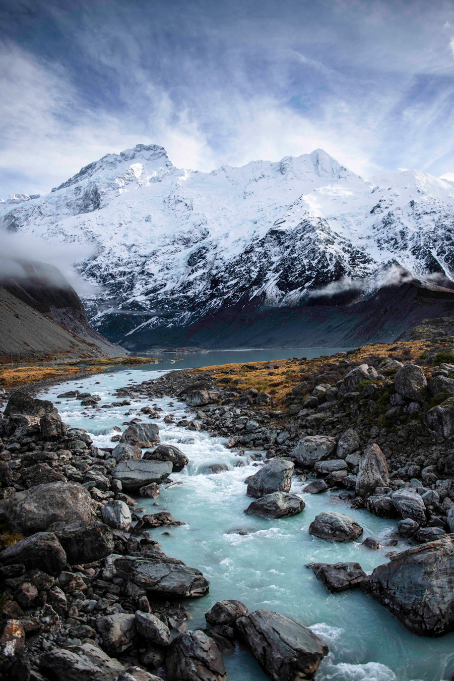 A turquoise river rushes through a rocky landscape towards a snow-capped mountain range under a cloudy sky. The foreground is filled with large, dark gray rocks, while the background features a mountain partially covered in snow and low-hanging clouds. Photo by Raina Wessen.