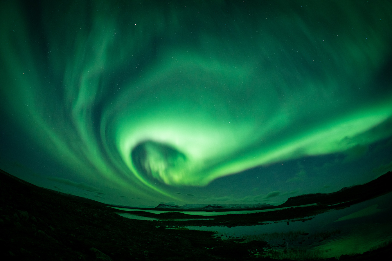 A wide-angle shot captures a vibrant green aurora borealis swirling dramatically across a starry night sky above a dark, rugged landscape with small bodies of water. The aurora's intense glow illuminates the scene, creating a mesmerizing display of natural light. Photo by Oscar Manguy.