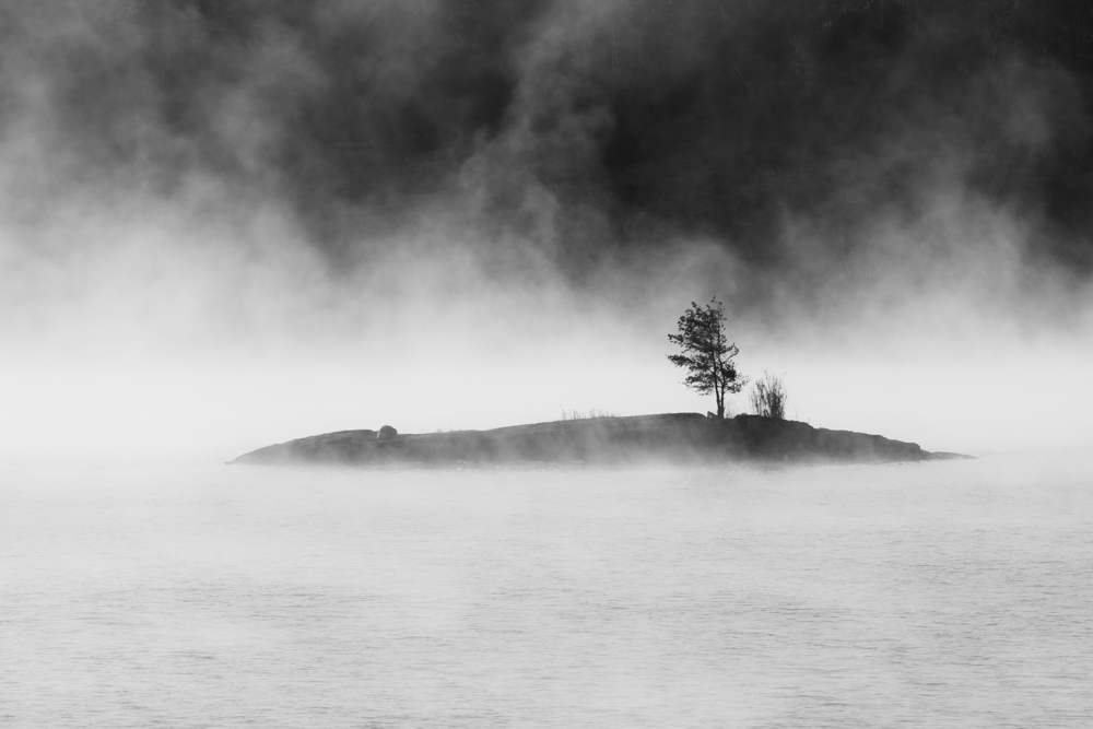 A black and white landscape photograph shows a small, dark island in the middle of a misty body of water, with a single tree standing on the island. The background is obscured by heavy fog or clouds, creating a sense of isolation and mystery. Photo by Brian Wessen.