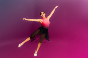 A smiling dancer in a pink and black leotard and black skirt leaps against a gradient purple background, arms outstretched and legs extended. She wears white ballet shoes. Photo by JSPS Schools.