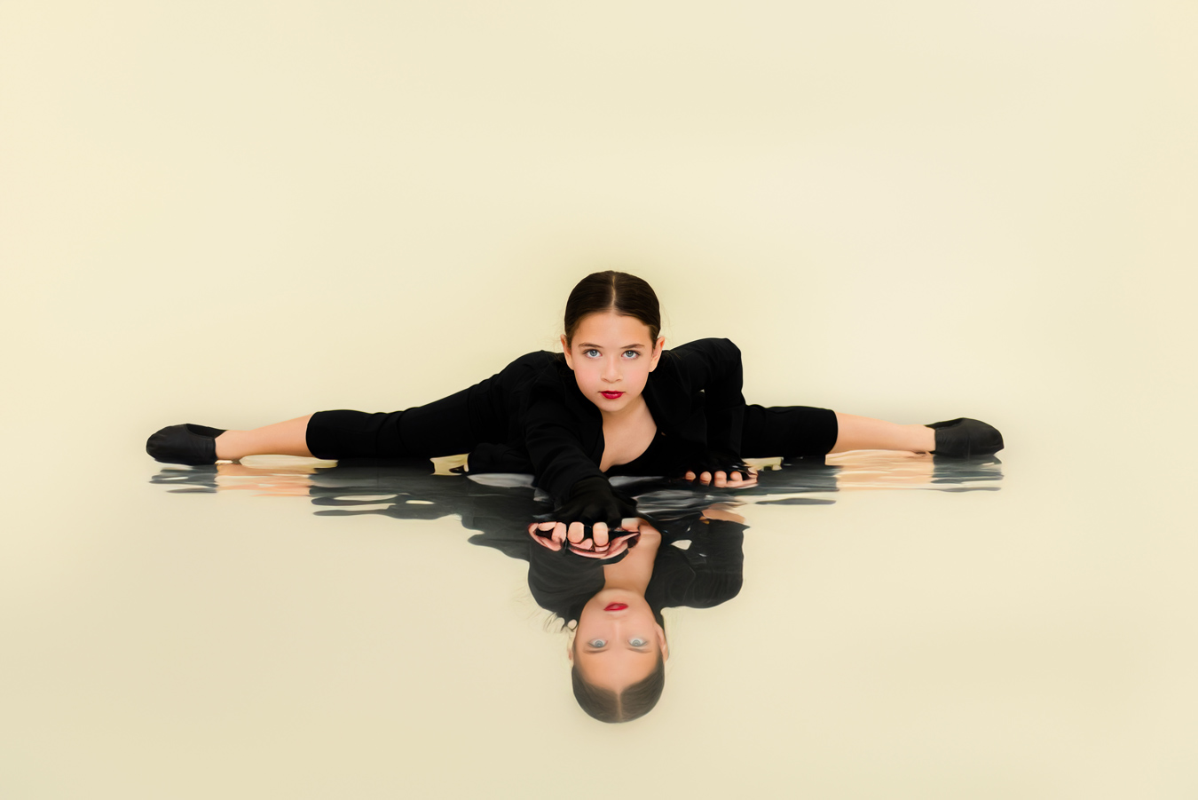 A young dancer in a black outfit and ballet shoes performs a split on a reflective surface, creating a mirrored image of her pose. She looks directly at the viewer with a focused expression. Photo by JSPS Schools.