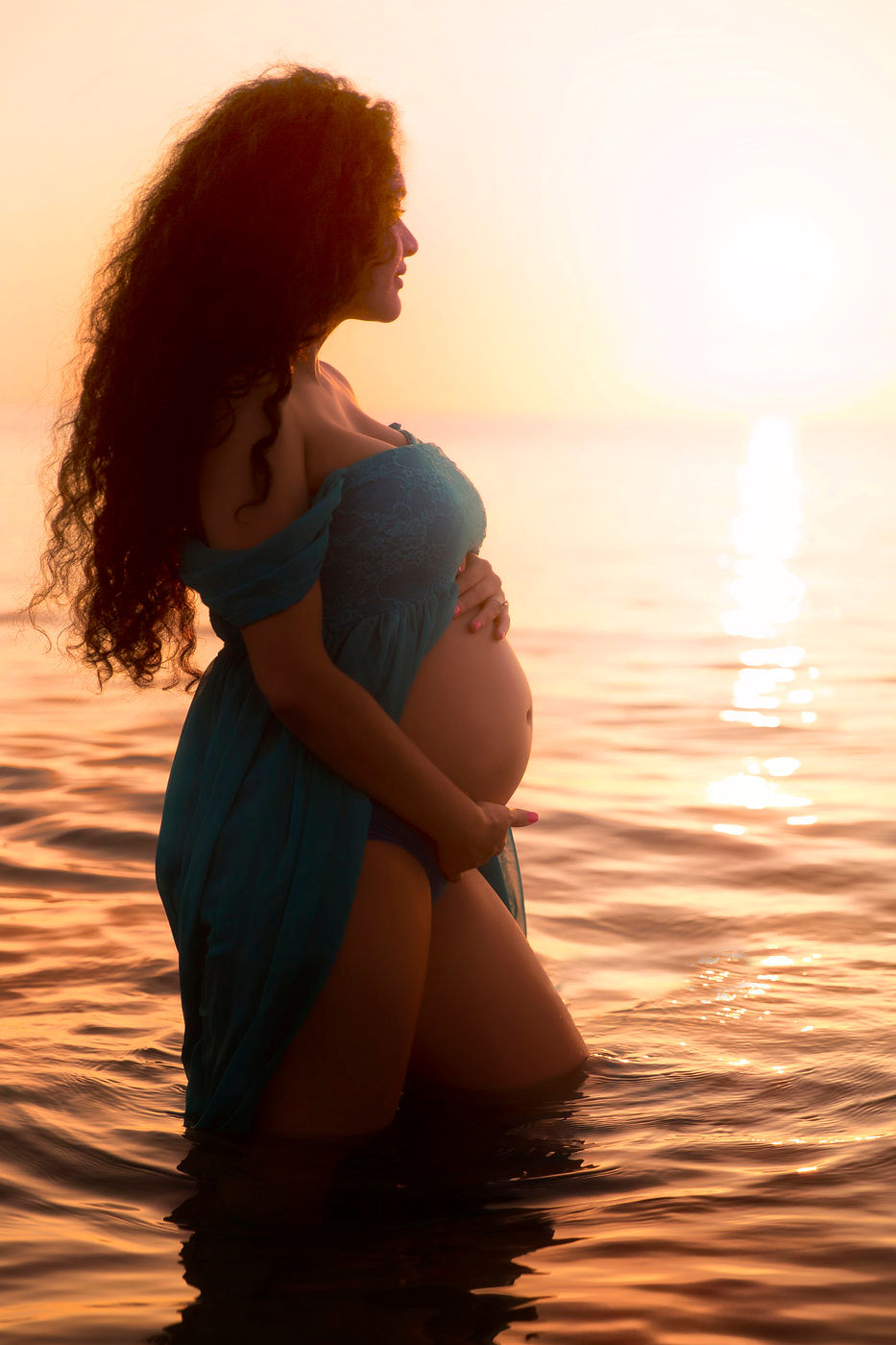 A pregnant woman with long, dark curly hair stands in the ocean at sunset, wearing a blue dress and cradling her baby bump. The golden light reflects off the water, creating a serene and beautiful scene. Photo by Adam Opris