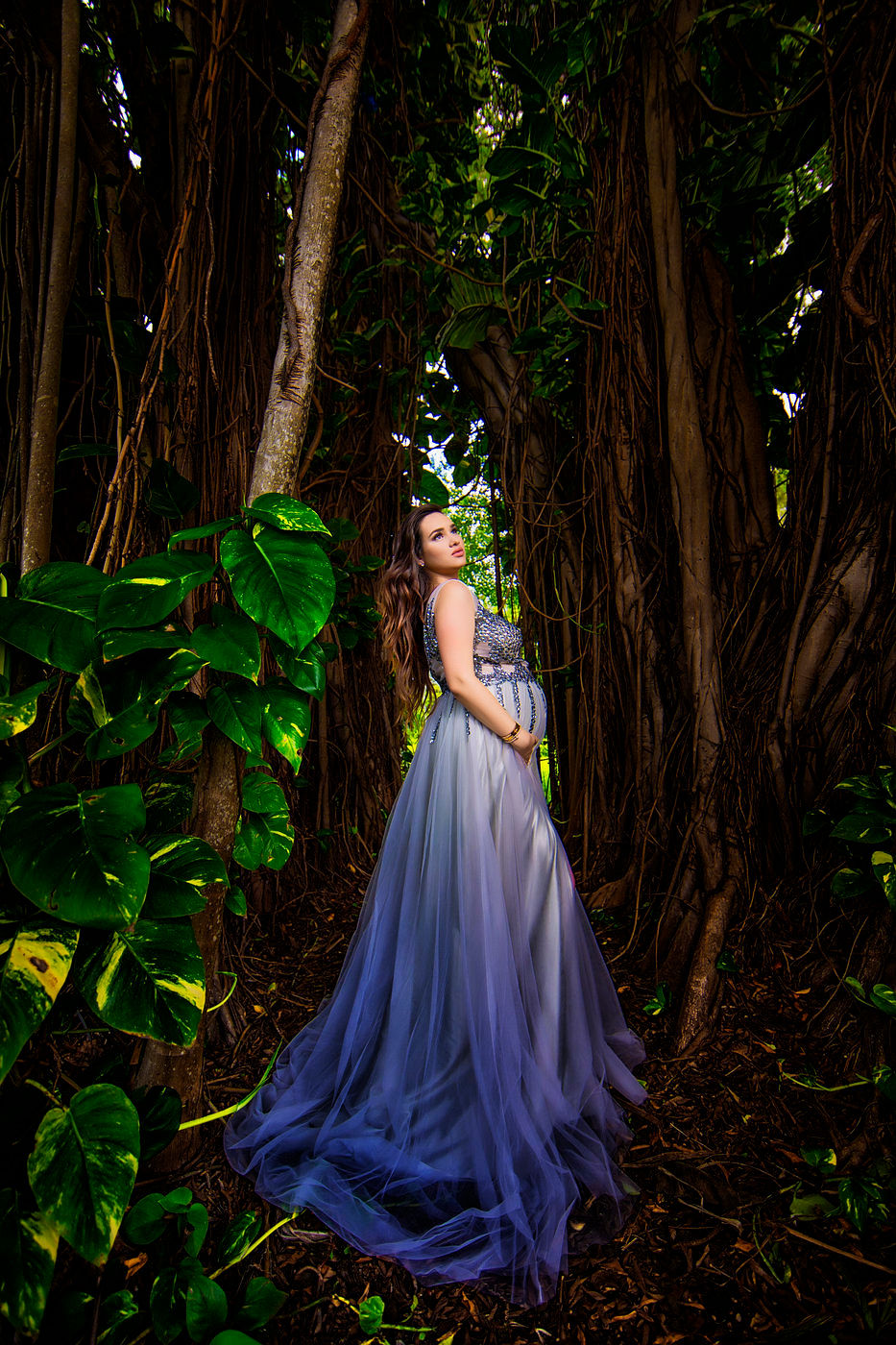 A pregnant woman in a flowing blue gown stands in a lush forest, cradling her baby bump and looking upwards. The scene is framed by large trees with hanging vines and vibrant green foliage. Photo by Adam Opris