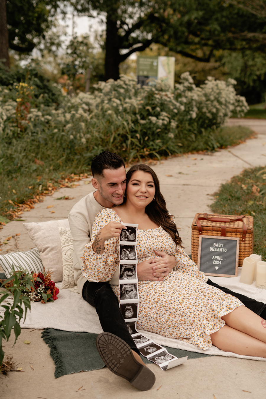 A smiling couple sits on a blanket outdoors, announcing their pregnancy; the woman, wearing a floral dress, holds a strip of ultrasound images, while a sign next to them reads "Baby DeSanto, April 2024." The scene is decorated with pillows, flowers, a picnic basket, and candles. Photo by Sam Reed.