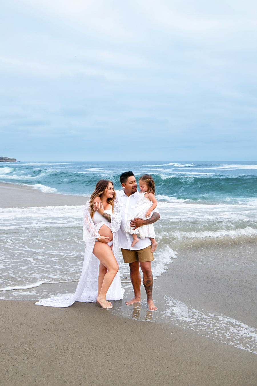 A family stands on a beach with waves crashing behind them; the pregnant mother wears a white swimsuit and lace cover-up, the father holds their young daughter in a white dress, and they all look at each other with smiles.