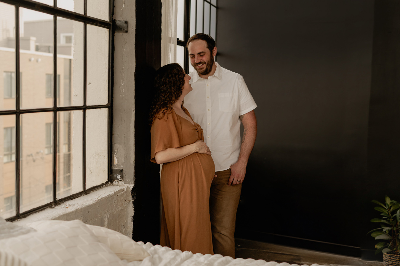 A pregnant woman in a brown dress stands by a window with her partner, who is smiling at her, in a warmly lit indoor setting. The couple is positioned near a large window with a view of a building, and a bed is partially visible in the foreground. Photo by Sam Reed.