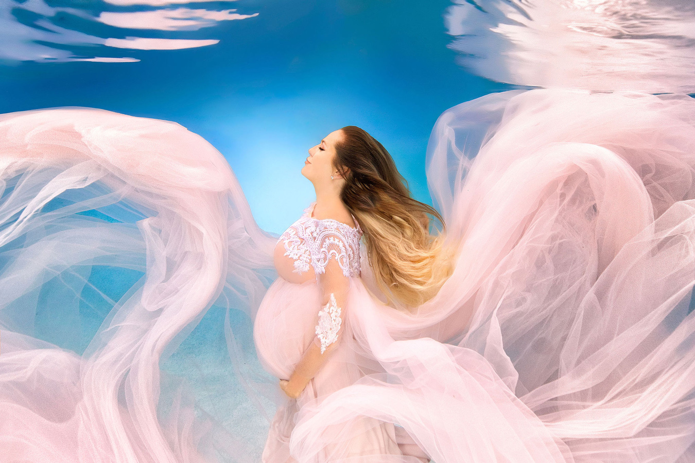Underwater, a pregnant woman in a flowing pink gown with lace details cradles her belly, her eyes closed and head tilted back against a bright blue background with water reflections above. The gown billows around her, creating a soft, ethereal effect. Photo by Adam Opris