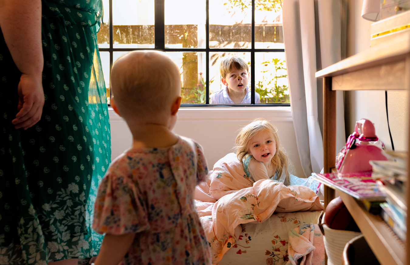 A young girl smiles from a low bed mattress inside a sunlit room while a toddler stands nearby and a young boy presses his face against the window from the outside.