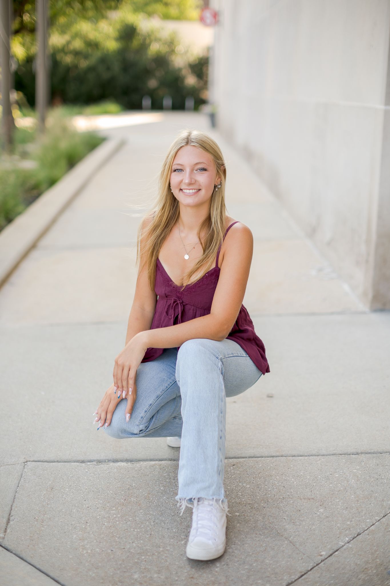Smiling young woman sitting outdoors on concrete steps wearing a burgundy top and light jeans