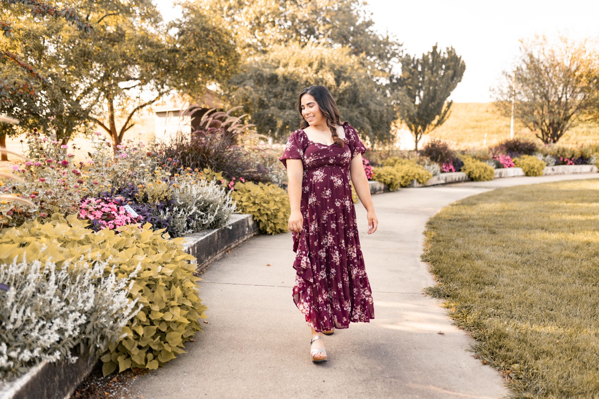 Woman in a burgundy floral dress walking along a garden path surrounded by blooming flowers