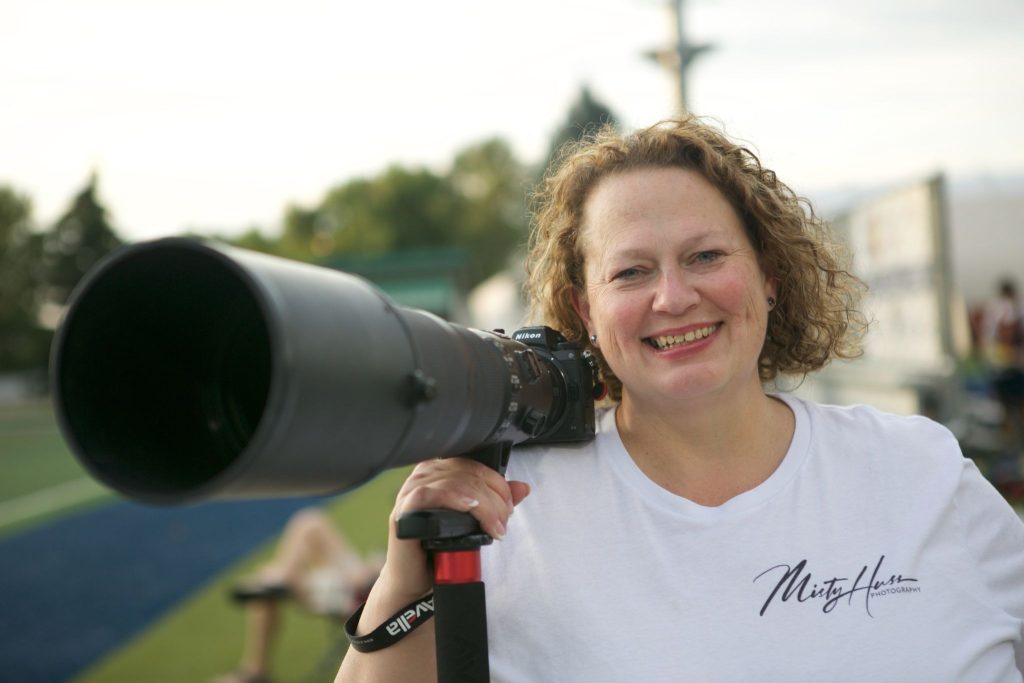 A smiling woman with curly blonde hair holds a professional Nikon camera with a massive telephoto lens resting on her shoulder while standing outdoors.